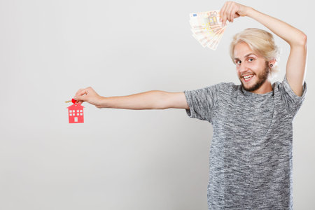 Household savings and finances, economy concept. Happy man holding money and keys to house, studio shot on grey backgroundの写真素材