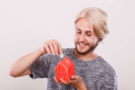Household savings and finances, economy concept. Smiling man putting money coin into piggy bank in shape of house, studio shot on light backgroundの写真素材