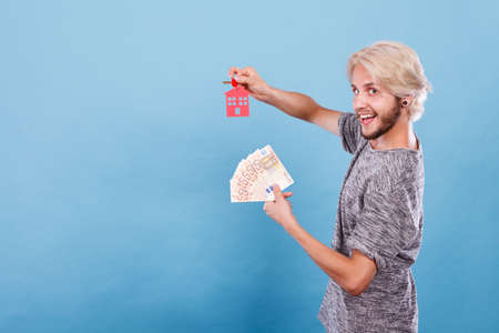Household savings and finances, economy concept. Happy man holding money and keys to house, studio shot on blue backgroundの写真素材