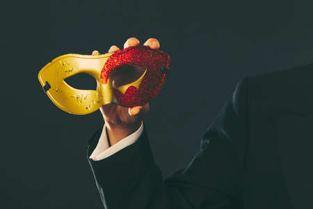 Holidays, people and celebration concept. Male hand holding red golden carnival mask closeup on darkの写真素材