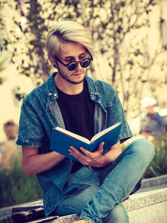 Male fashion, student concept. Guy holding and studying from notebook wearing jeans outfit and eccentric sunglasses sitting on white ledgeの写真素材