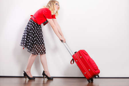 Traveling vacation concept. Elegant young woman in full length wearing polka dot black dress pulling heavy red travel bagの写真素材