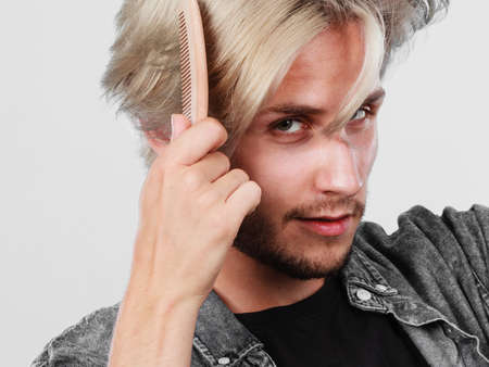 Hairdo coiffure hairstyle concept. Handsome young fashion model combing his colored blonde hair using wooden comb tool, stylish haircutの写真素材
