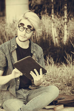 Male fashion, technology, student concept. Guy with tablet wearing jeans outfit and eccentric sunglasses sitting on white ledgeの写真素材