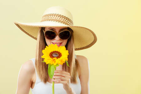 Closeup of attractive summer woman in sunglasses straw hat with sunflower in her hand on yellow backgroundの写真素材