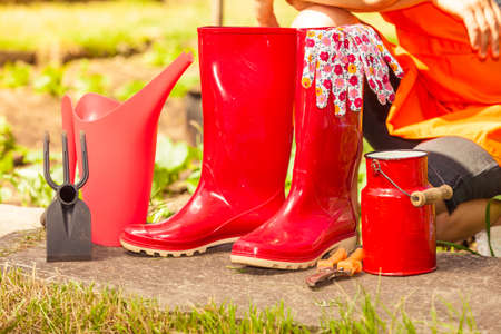 Gardening tools outdoor in garden, red rubber boots and water can. Woman farmer in hat in the backgroundの写真素材