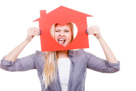Ownership and property concept. Happy woman holding red paper house with hole in heart shape, loving new home, folling around and sticking tongue outの写真素材