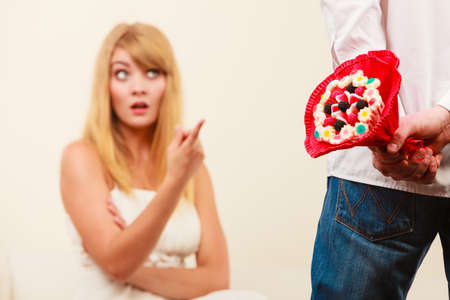 Man with candy bunch flowers behind back and pretty unhappy bored woman. Boyfriend with present gift trying to apologize girlfriend.の写真素材