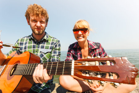 Happy young man hipster playing guitar for woman by sea ocean. Handsome guy and pretty girl in...