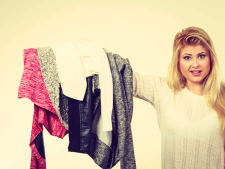 Fashion, clothes dilemmas concept. Young blonde woman teen girl holding big pile of warm winter clothing, cant decide what to wear, toned imageの写真素材