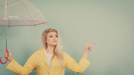 Blonde woman wearing yellow raincoat holding transparent umbrella checking weather if it is raining...