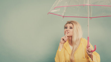 Blonde woman wearing yellow raincoat holding transparent umbrella checking weather if it is raining showing silence gesture.の写真素材