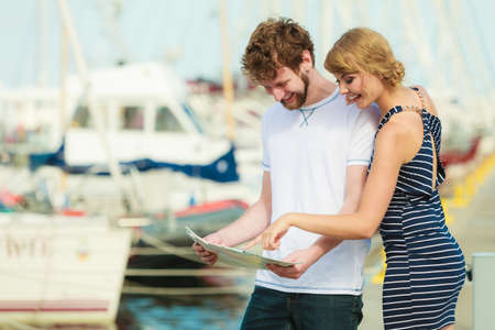 Summer travel concept. Young tourist couple on vacation standing in front of boats in marina looking up directions on mapの写真素材