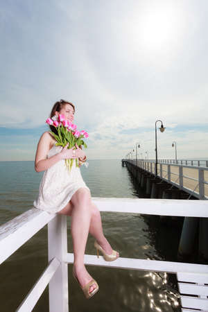 Feminity, bridesmaid, travel concept. Beautiful woman wearing white dress holding a bouquet of pink flowers sitting on wooden pierの写真素材