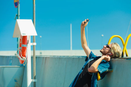 Contacts, technology, self-esteem concept. Young blonde man on vacations using his smartphone to take cool selfie shotの写真素材