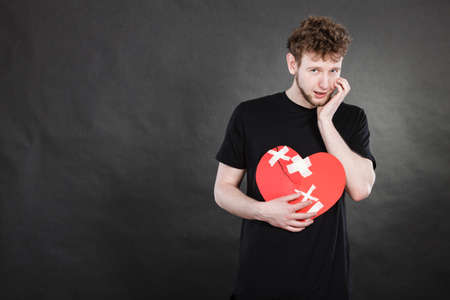 Bad relationships, breaking up, sadness emotions concept. Very sad young man holding broken heart made of paper.の写真素材