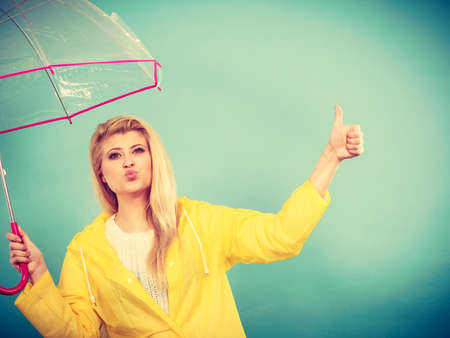 Blonde woman wearing yellow raincoat holding transparent umbrella checking weather if it is raining showing thumb up gestureの写真素材