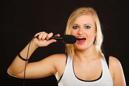 Karaoke, music, singer concept. Portrait of blonde woman singing to microphone, young star performing, studio shot, black background.の写真素材