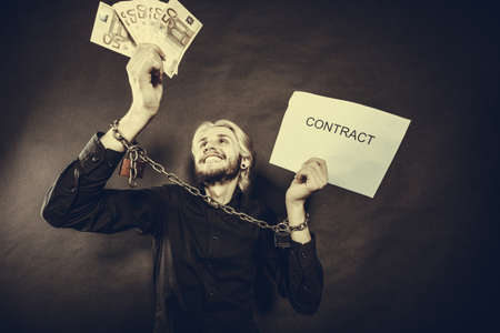 Stress at work, no freedom, pursuit of money concept. Man with chained hands choosing between money and contract, studio shot on dark, grunge backgroundの写真素材
