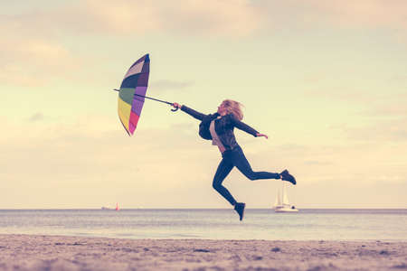 Happiness, enjoying cold autumn weather, feeling great concept. Woman jumping with colorful umbrella on beach near sea, sunny day and clear blue skyの写真素材