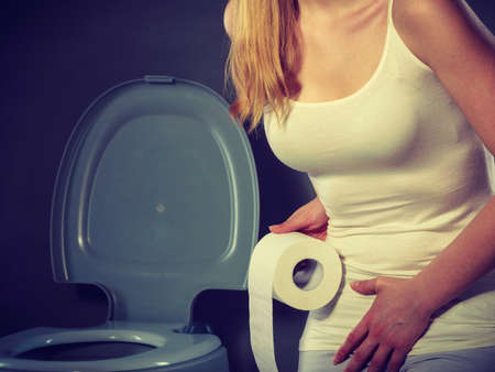 Sick woman with hands pressing her crotch lower abdomen, holding paper roll in front of toilet bowl. Medical problems, incontinence, health care conceptの写真素材