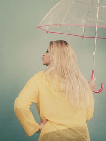 Rainy autumn day accessories ideas concept. Blonde woman wearing raincoat holding clear transparent umbrella, back view.の写真素材
