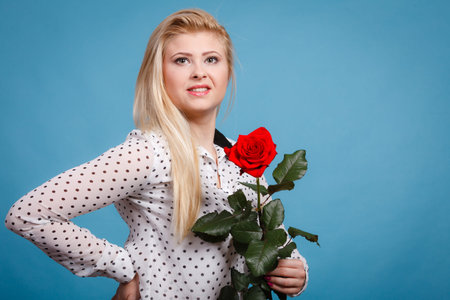 Woman holding red rose. Lovely blonde smiling girl with flower studio shot on blue. Romance, holidays, valentine day conceptの写真素材