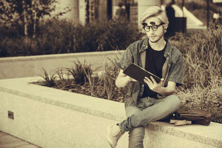 Male fashion, technology, student concept. Guy with tablet and bag full of notebooks wearing jeans outfit sitting on white ledge next to flowers, sepiaの写真素材