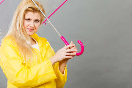 Good mood during rainy day. Happy blonde woman wearing yellow raincoat holding transparent umbrellaの写真素材