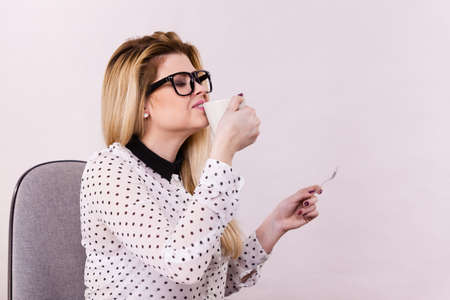 Happy young woman wearing eyeglasses at office drinking hot coffee or tea enjoying her break time during work. Job relax.の写真素材