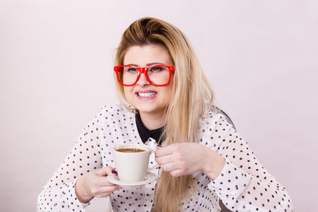 Happy young woman wearing eyeglasses at office drinking hot coffee or tea enjoying her break time...