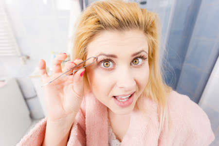 Woman in bathroom plucking eyebrows depilating with tweezers. Girl tweezing removing her facial hairs. Unusual wide angle viewの写真素材