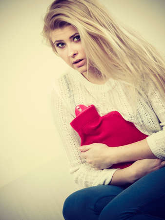 Painful periods and menstrual cramp problems concept. Woman having stomach cramps sitting on cofa feeling very unwell holding hot water bottle to feel some reliefの写真素材