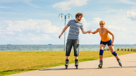 Holidays, active people and friendship concept. Young fit couple on roller skates riding outdoors on sea coast, woman and man rollerblading together on the promenadeの写真素材