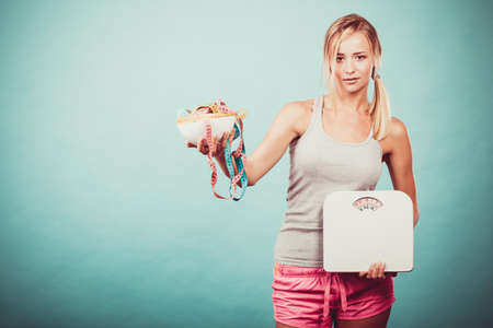 Diet, healthy eating and slim body concept. Fit fitness girl holding bowl with many colorful measuring tapes as dieting symbol and weight scales studio shot on blueの写真素材