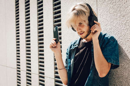 Men fashion, technology, urban style clothing concept. Hipster guy standing on city street wearing jeans outfit and sunglasses listening to music and holding phoneの写真素材