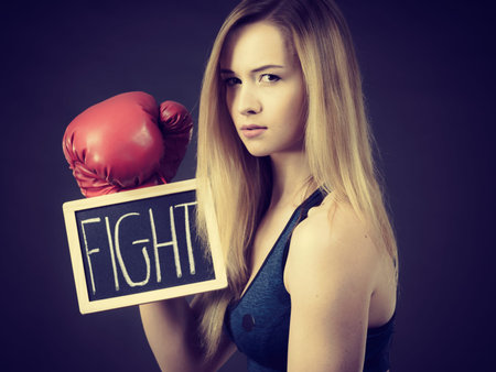Young sporty woman wearing boxing glove holding black board with fight text sign. Studio shot on...