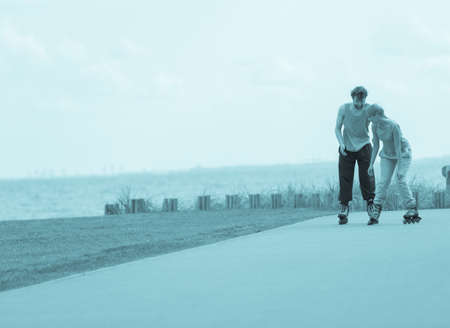 Active holidays, exercises, relationship concept. Young woman and man dressed up in sporty way, holding their hands while rollerblading together on promenade, blue colorsの写真素材