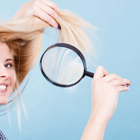 Haircare and hairstyling, bad effects of bleaching concept. Happy blonde woman looking at her damaged, split hair ends through magnifying glass.の写真素材