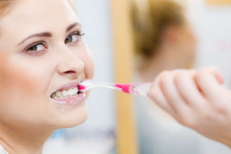 Woman brushing cleaning teeth closeup. Blonde girl with toothbrush in bathroom, looking at mirror. Oral hygiene.の写真素材
