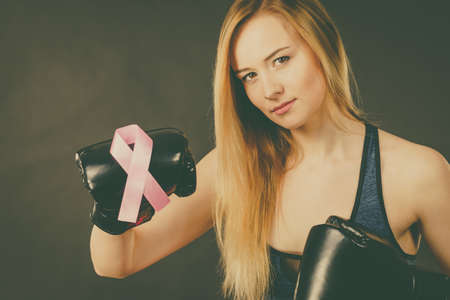 Young sporty woman wearing boxing gloves having pink ribbon tape, breast cancer symbol. Fighting with disease, feminine motivation. Studio shot on dark background.の写真素材