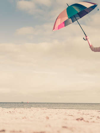 Weather, rain protection concept. Woman hand holding colorful umbrella on beach, sunny day and clear blue skyの写真素材