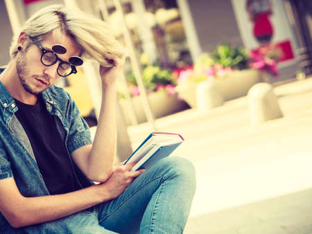 Male fashion, student concept. Guy holding notebook wearing jeans outfit and eccentric sunglasses sitting on white ledge next to modern buildingの写真素材