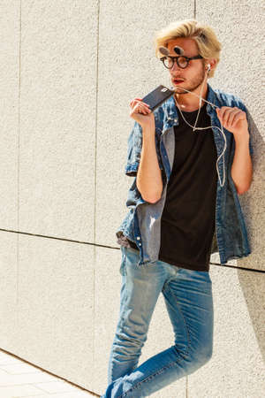 Men fashion, technology, urban style clothing concept. Hipster guy standing on city street wearing jeans outfit and eccentric sunglasses listening to music and holding phoneの写真素材