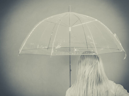 Rainy autumn day accessories ideas concept. Blonde woman wearing raincoat holding clear transparent umbrella, back view.の写真素材