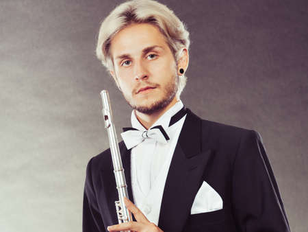 Classical music, passion and hobby concept. Portrait of elegantly dressed musician blonde young man holding flute. Studio shot on dark grey backgroundの写真素材