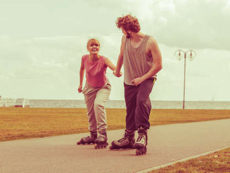 Active holidays, exercises, relationship concept. Young man dressed in sports clothes putting his girlfriend up to do rollerblading while holding her hand on promenadeの写真素材