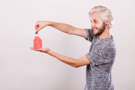 Household savings and finances, economy concept. Man putting money coin into piggy bank in shape of house, studio shot on light backgroundの写真素材