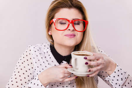 Happy young woman wearing eyeglasses at office drinking hot coffee or tea enjoying her break time during work. Job relax.の写真素材