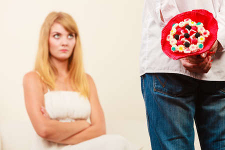 Man with candy bunch flowers behind back and pretty unhappy bored woman. Boyfriend with present gift trying to apologize girlfriend.の写真素材